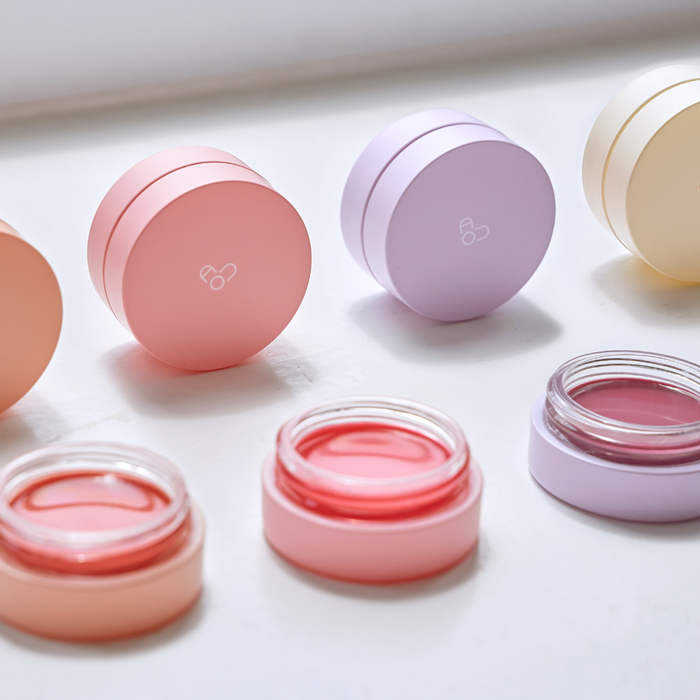 AOU Glowy Tint Balm jars in various pastel colors on white surface, showcasing the product's attractive packaging and shades.