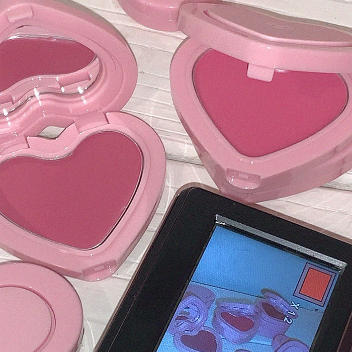 Heart-shaped pink AOU Soft Cream Blush in open compact cases next to a digital display on a wooden surface.