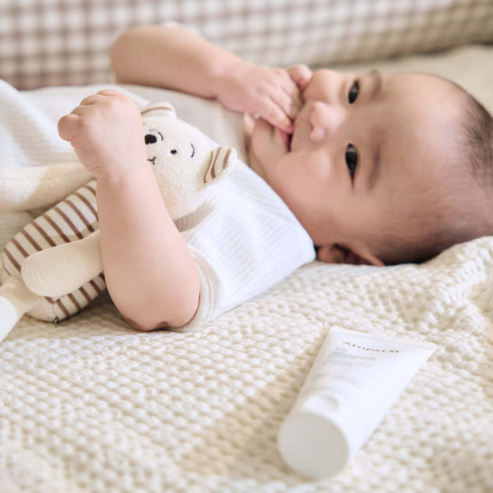 Baby lying on a blanket with stuffed toy next to ATOPALM Panthenol Lotion 180ml tube.