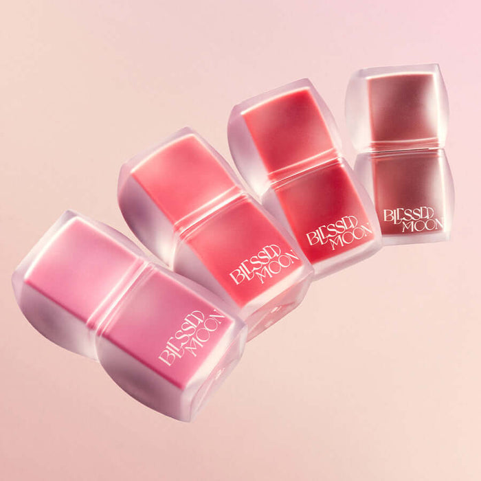 BLESSED MOON Veil Blur Lip Tint in various shades on a pink background, four lip tints lined up with sleek packaging.