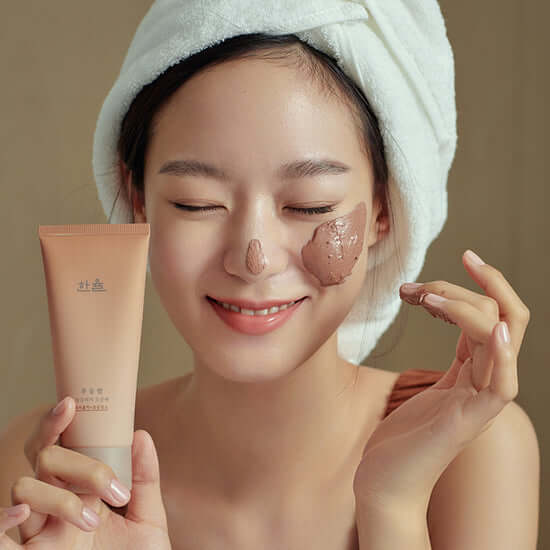 Woman applying HANYUL Buddle Night Cleansing Pore Pad with a smile, wearing a towel on her head, emphasizing skincare routine.