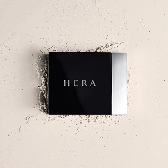 Close-up of HERA Airy Powder Primer 8.5g on a textured surface, showcasing sleek packaging and brand name.