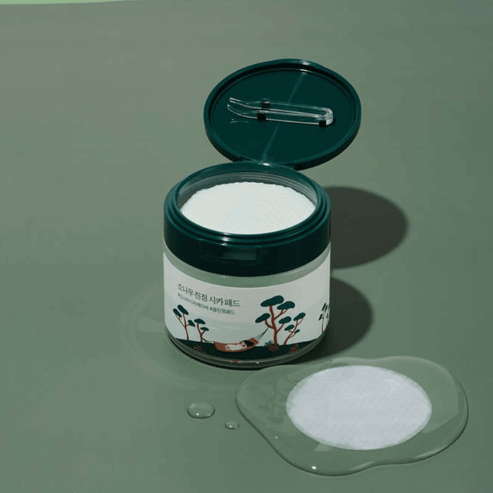 Open jar of ROUND LAB Pine Calming Cica Pads 50 sheets with one pad and tweezers on a green surface.