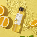 SKINFOOD Yuja C Dark Spot Clear Toner 200ml bottle surrounded by sliced yuja fruit and clear water ripples on a sunny yellow background.