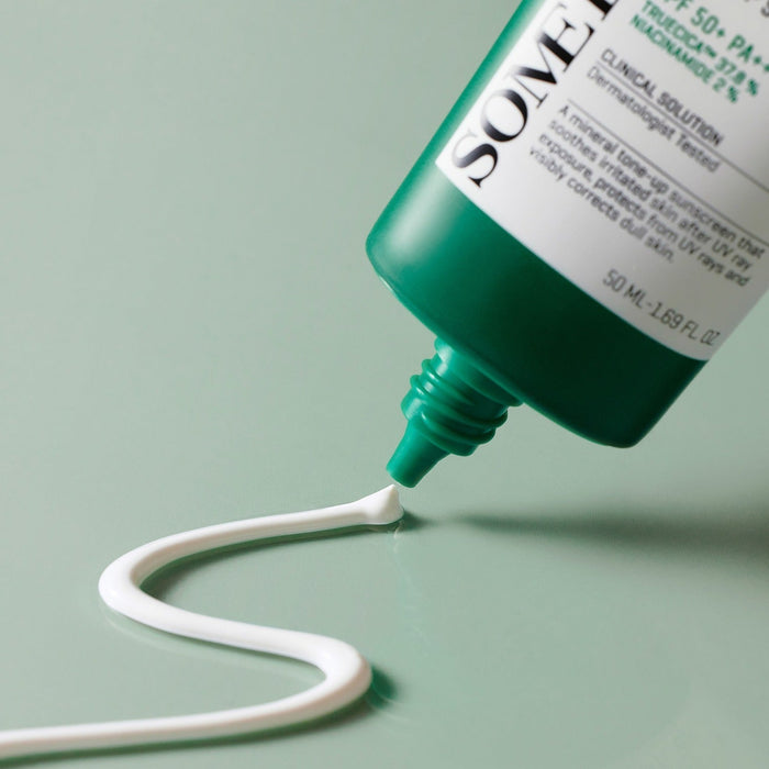 SOME BY MI Truecica Weaponless Calming Tone Up Sun Cream 50ml being dispensed from tube, showcasing texture and color on green surface.