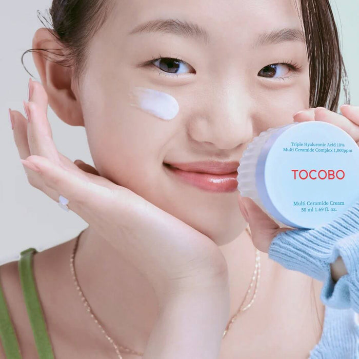 Smiling person applying TOCOBO Multi Caramide Cream to their face, holding a jar of the cream, showcasing skincare routine.