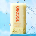 TOCOBO Vita Waterproof Sun Stick SPF50+ PA++++ with water droplets against a blue sky background.