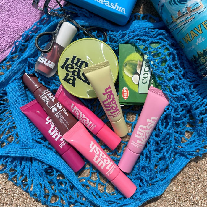 Various UNLEASHIA products including the UNLEASHIA Sunset Dazzle Gloss Balm displayed on a blue net.